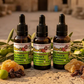 Three bottles of Anointing Oil product with green labels on a textured surface with leaves and herbs.