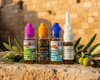 Four small bottles of hair growth and conditioning liquid, including Vela Skin Vela-Science and Vela-Organics Colof 100% Olive Hair Growth & Conditioning liquids, arranged on a stone ledge with olive branches in front of a warm stone wall background.