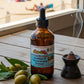 Bottle of healing oil with olives and a smoking device on a wooden surface with a beach view.