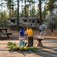 Two bottles of oil, one labeled 'Rx 9' and the other 'Oil of Elijah', on a picnic table and a campground setting with an RV and a forested state park with campfire burning. Around the bottle and at the base are olives and olive leaves and it incense burner with smoke rising from it to the right.